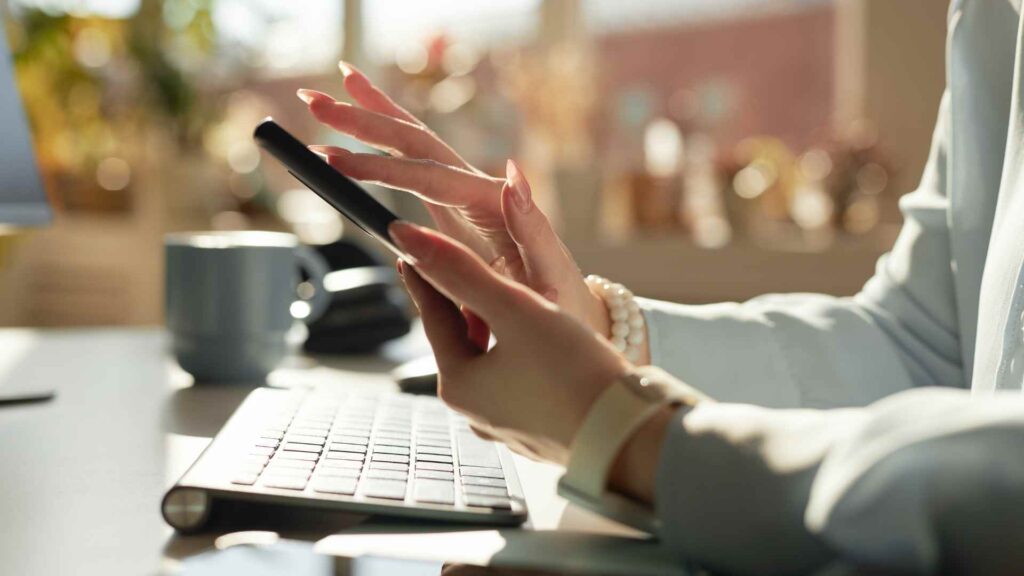 A person sitting at a desk using a smartphone next to a keyboard and a mug