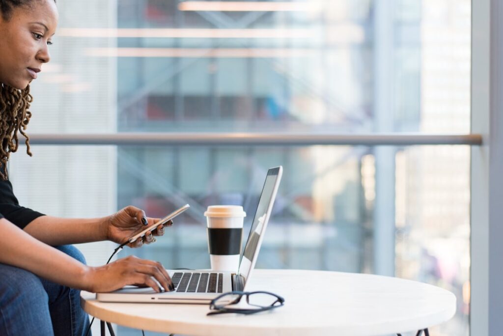 Woman working on a laptop at a small table while holding a smartphone, with a takeaway coffee nearby in a modern indoor space.