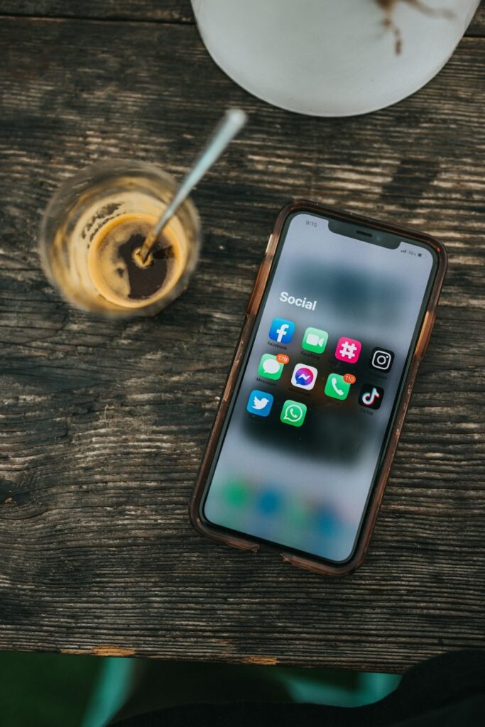 Smartphone lying on a wooden table displaying social media app icons next to a glass of iced coffee.