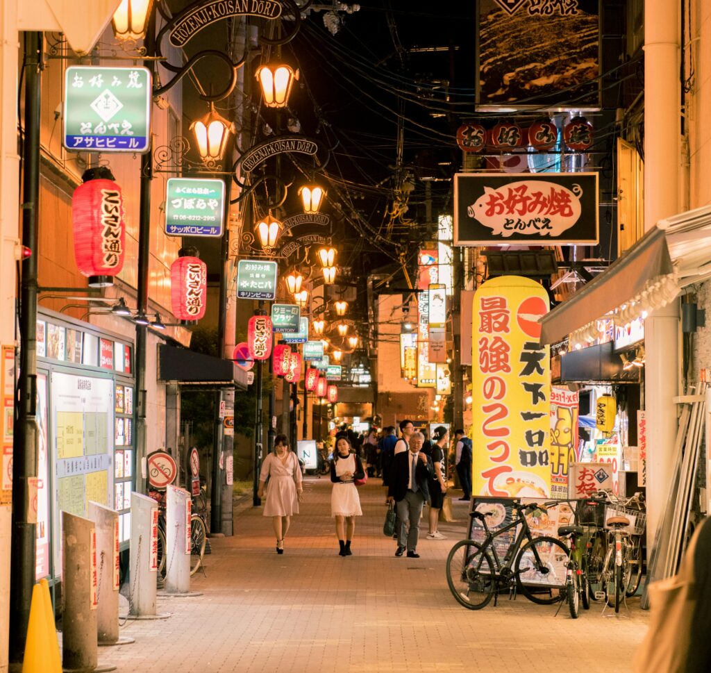 Nighttime street in Japan with illuminated lanterns, colorful shop signs, bicycles, and pedestrians walking through a lively alley