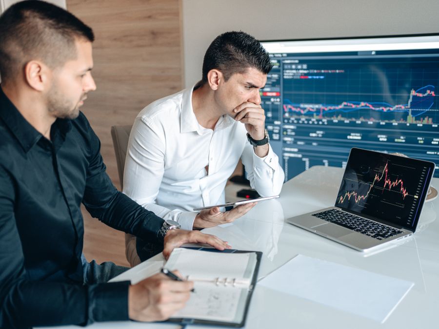 Two businessmen analyzing stock market data on laptops and tablets in an office setting.