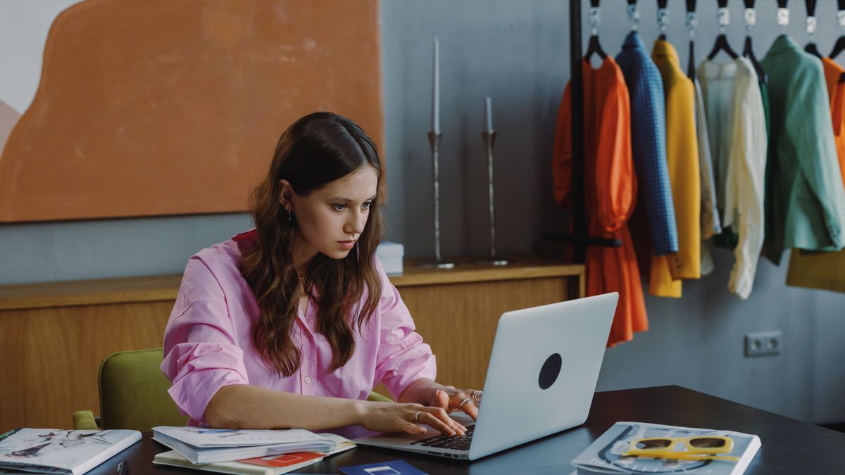 Businesswoman working at laptop in stylish office setting with clothes rack and design materials.