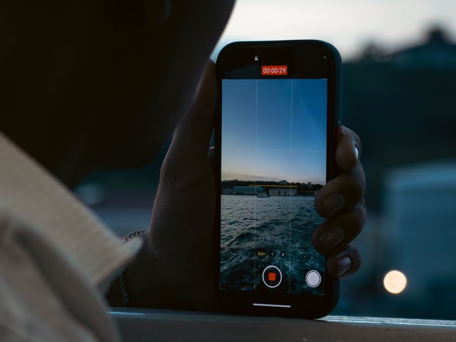 A person records a peaceful waterfront scene using a smartphone at dusk.