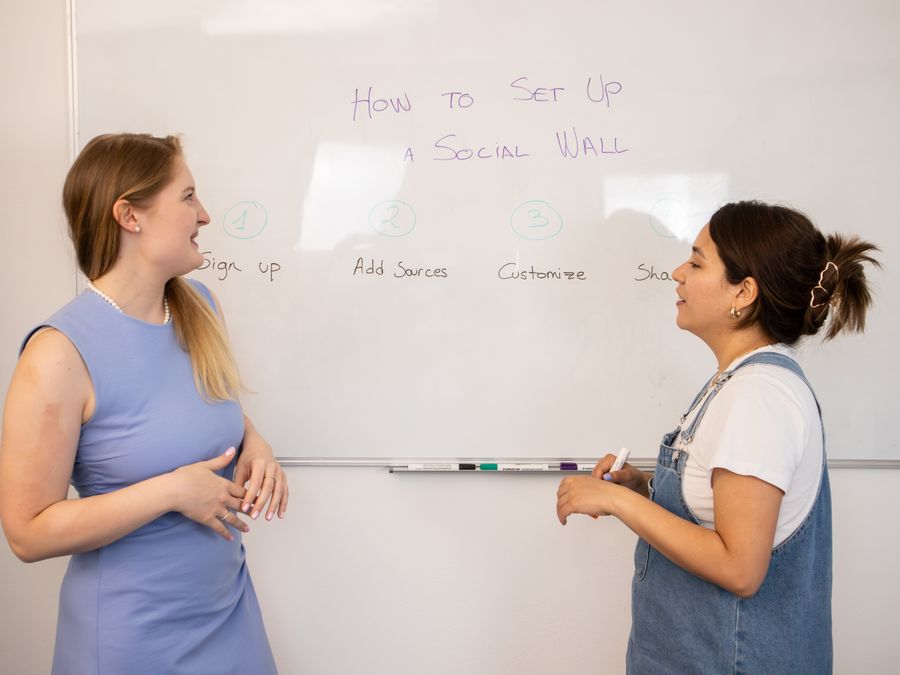 Two women discussing social media wall setup with tips on a whiteboard.