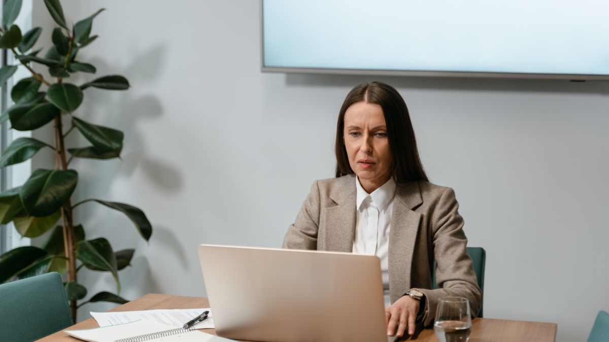 A woman in a business suit using a laptop at an office desk with documents and an indoor plant.