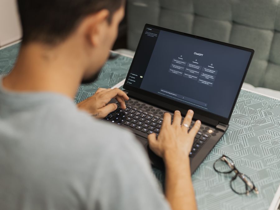 Man using laptop with AI interface, typing with focused attention. Indoors with eyewear beside.