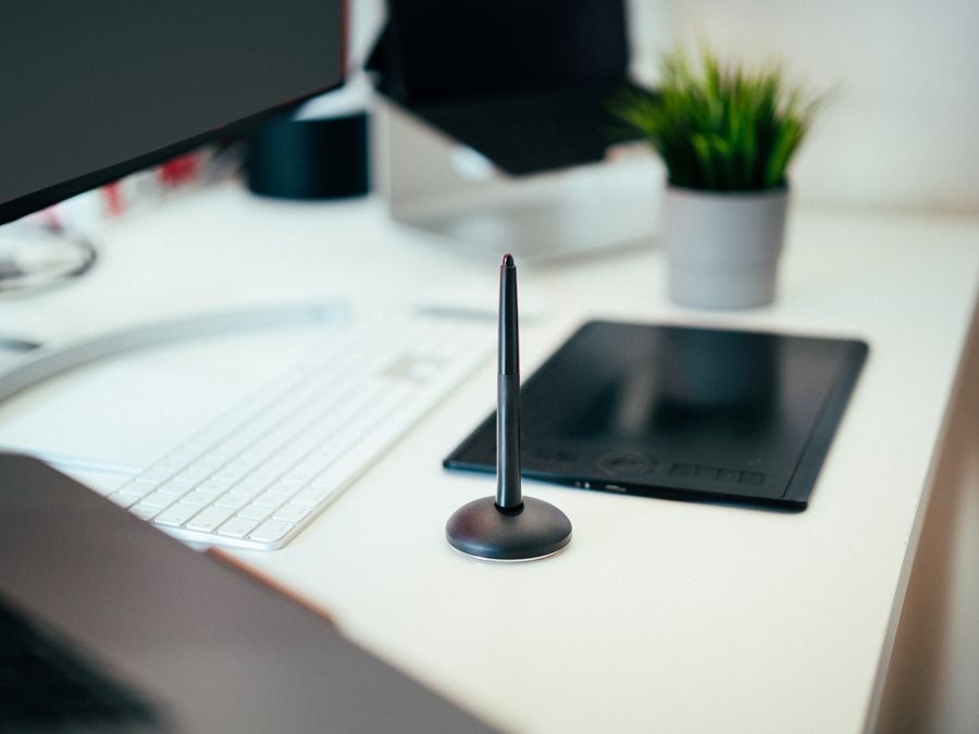 A modern workspace featuring a tablet, stylus, keyboard, monitor, and potted plant.