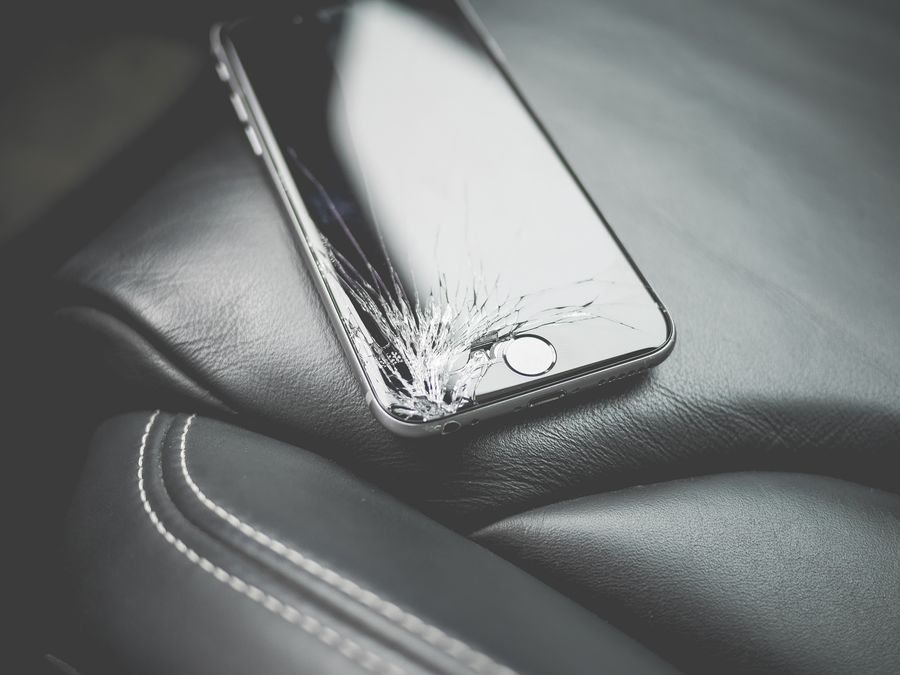 Close-up of a broken smartphone with a cracked screen on a leather car seat.