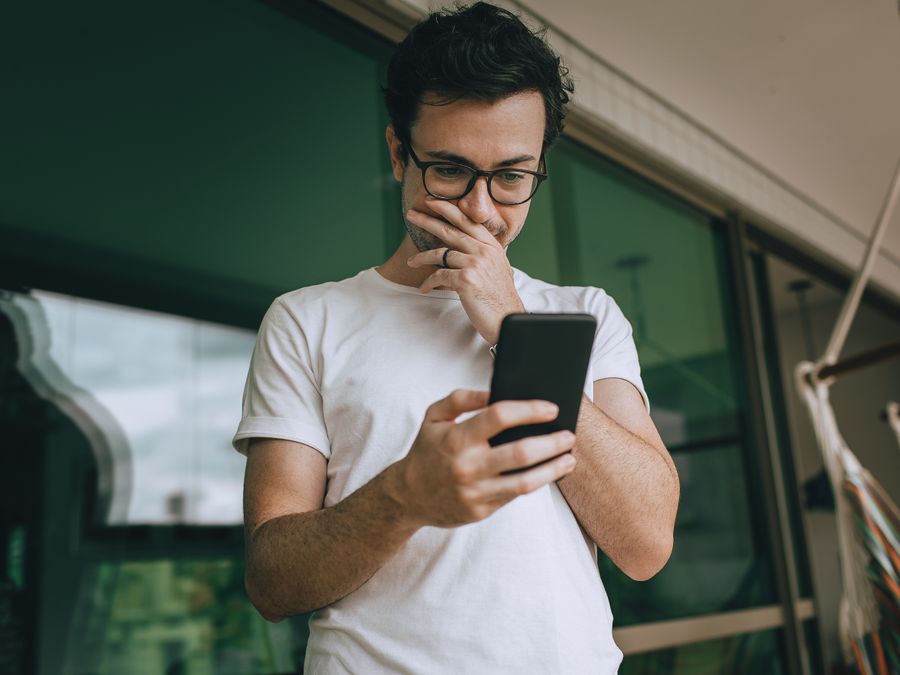 A young man in glasses standing on a balcony, using his smartphone with a thoughtful expression.