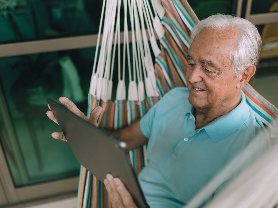 Senior man relaxing in a hammock indoors while using a tablet, enjoying leisure time.