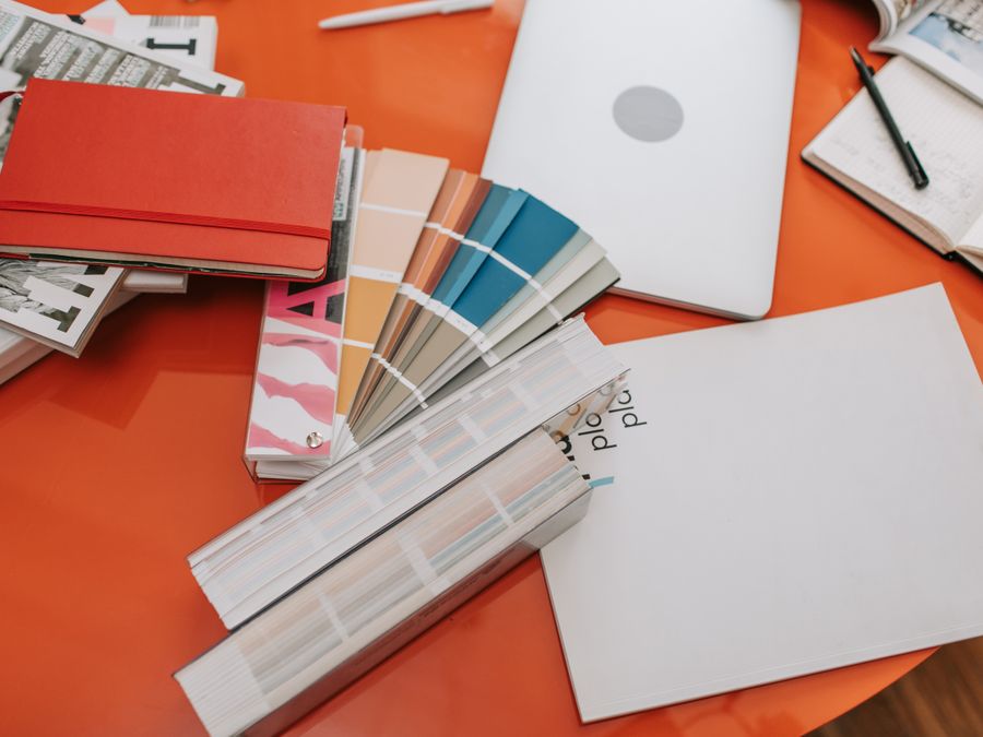 A vibrant workspace featuring design swatches, books, and a laptop on a bright orange desk.