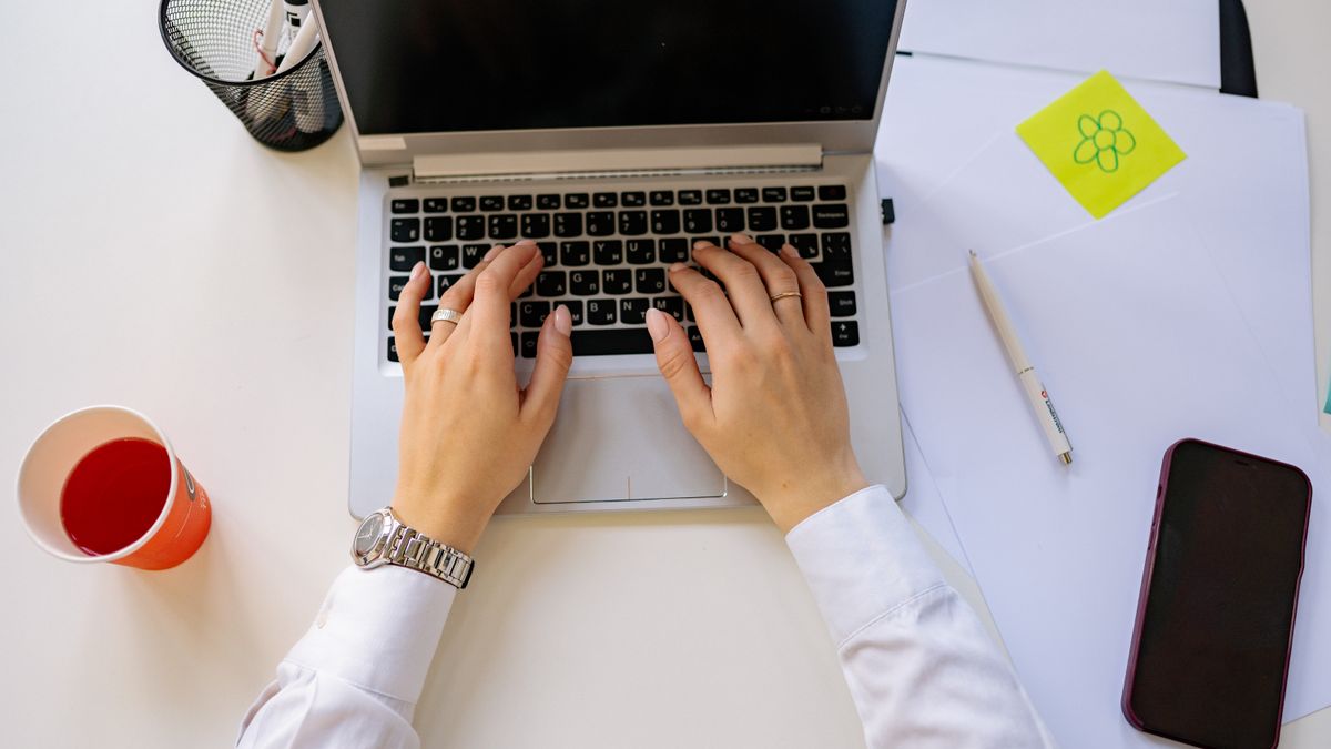 A person working on a laptop in a bright workspace with a drink and smartphone nearby.