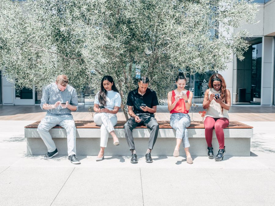 Five diverse people sitting on a bench outdoors, engaged with their smartphones.