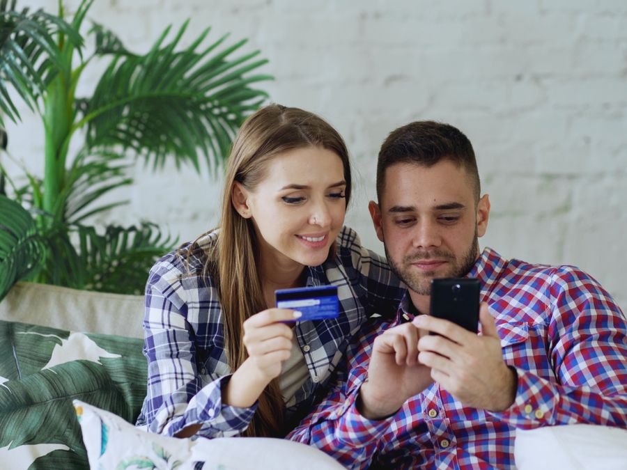 Smiling couple sitting on a sofa, shopping online with a smartphone and credit card.