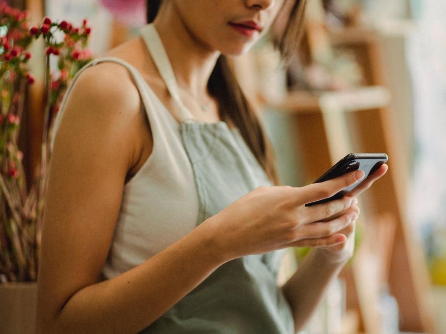 Crop anonymous florist in apron browsing mobile phone while working in cozy floral shop