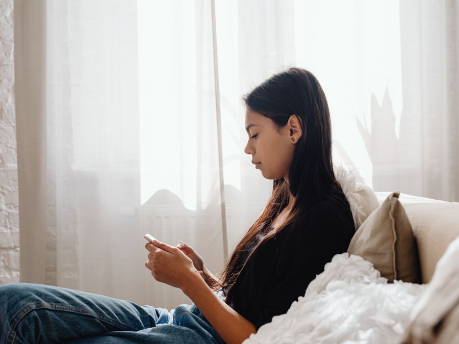 A teenager sits comfortably on a couch, focused on her smartphone in a peaceful indoor setting.