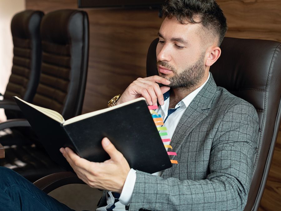 Businessman in suit reading notebook in modern office setting, planning and strategizing.
