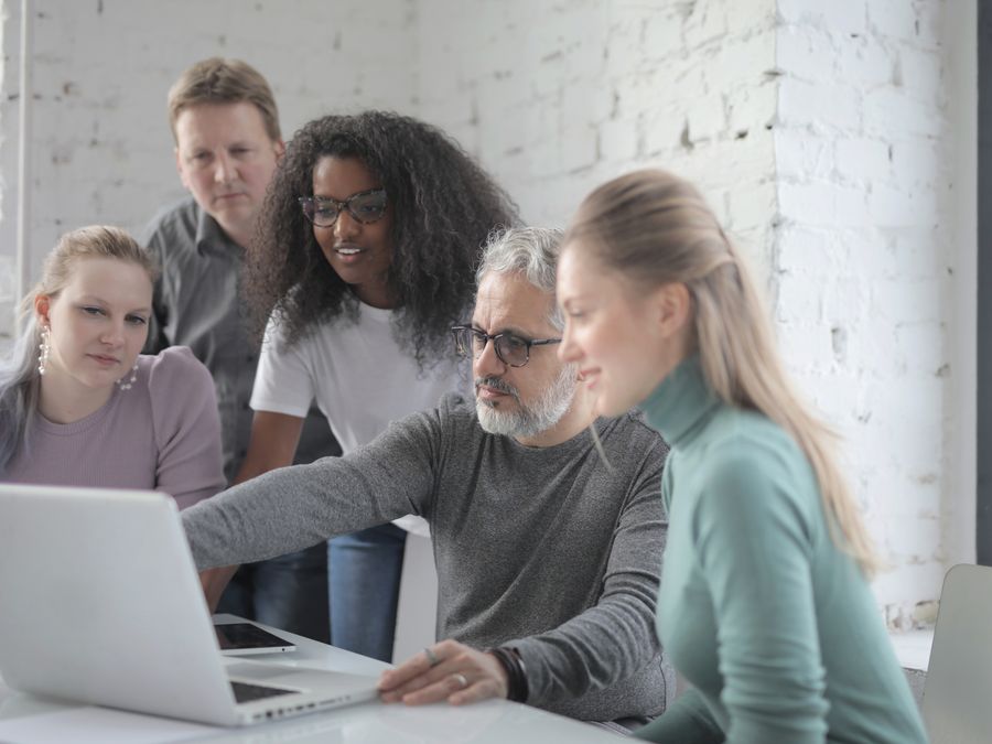 Group of diverse colleagues sitting and standing near table with laptop while discussing project and working together during business meeting