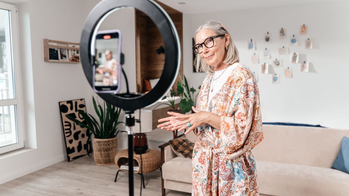 Elderly woman filming social media video with smartphone and ring light in modern living room.