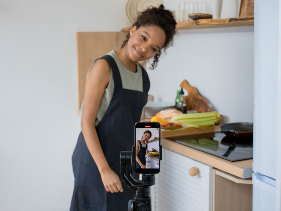 Smiling young woman films a cooking video at home using a smartphone in her kitchen.
