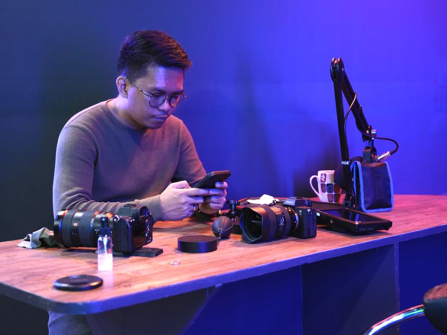 Man sitting at a desk with cameras and microphone, using smartphone.