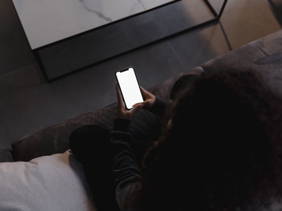 A person holding a smartphone with a blank screen, seated indoors on a couch.