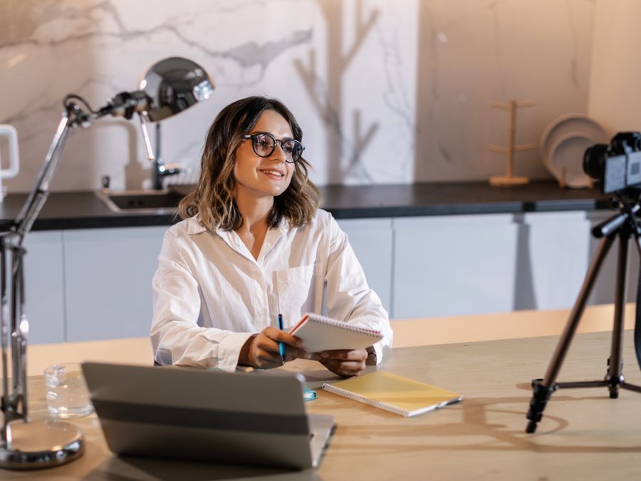 Young woman in glasses working indoors with laptop and camera, smiling while taking notes.