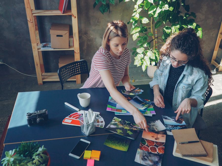 Two women collaborate on a creative project, analyzing photos and color palettes in a modern office.