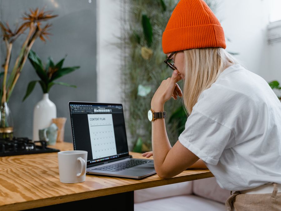 Young woman in orange beanie working on a laptop with a content plan in a cozy home office setting.