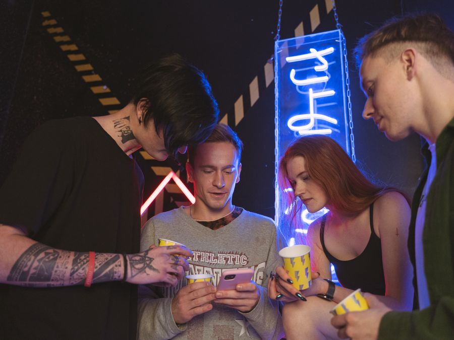 Group of young adults socializing indoors with neon lights, using smartphones.
