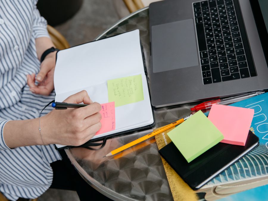 Person writing in notebook with laptop and sticky notes on a desk. Creative and organized workspace.