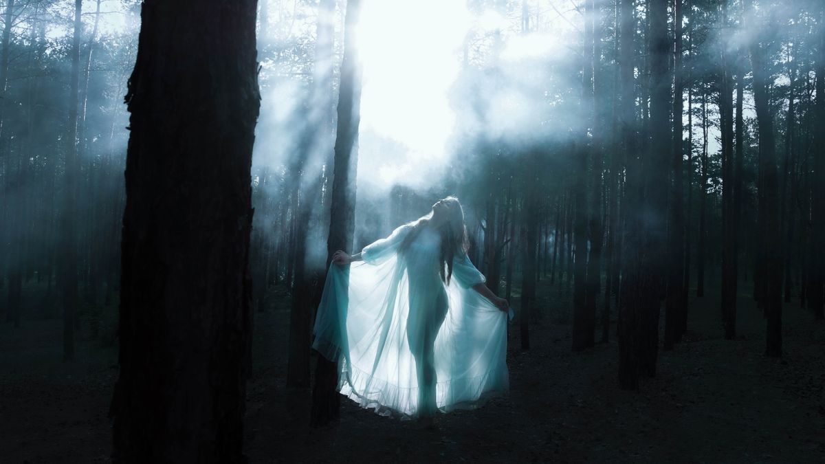 A woman in a flowing dress stands in a mystical forest with sunlight rays breaking through.