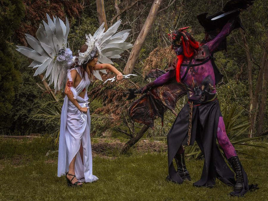 An epic battle between angel and demon cosplayers with costumes and props outdoors in a lush forest.