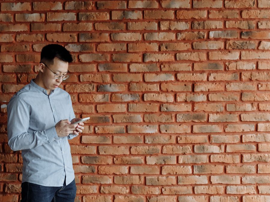Asian man texting on smartphone while standing against a brick wall, looking engaged.