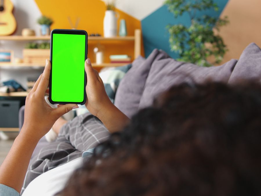 Person holding smartphone with green screen while relaxing on a couch indoors.