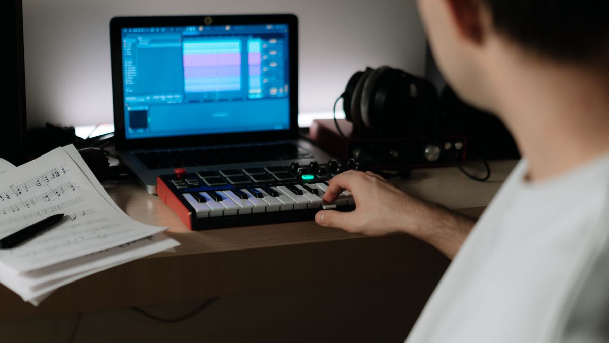 A music producer uses a synthesizer and laptop for composing in a home studio.