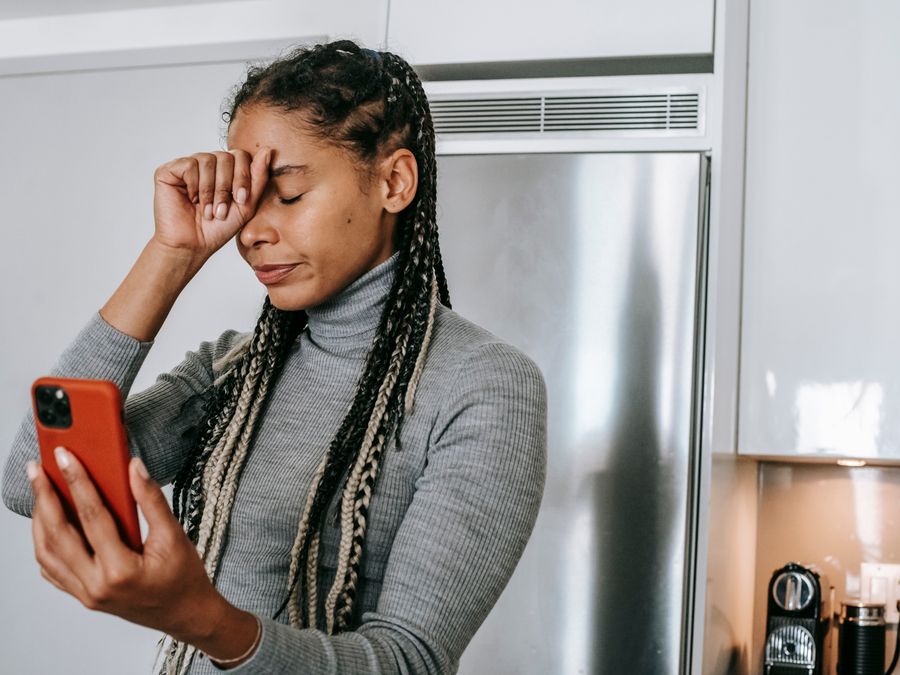 Unhappy young African American female with long braids in turtleneck touching face with closed eyes while having video conversation on smartphone in kitchen