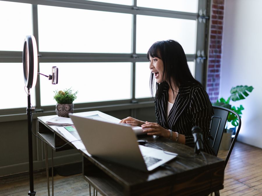 Asian woman in office setting recording content with laptop and ring light, showcasing elegant business setup.