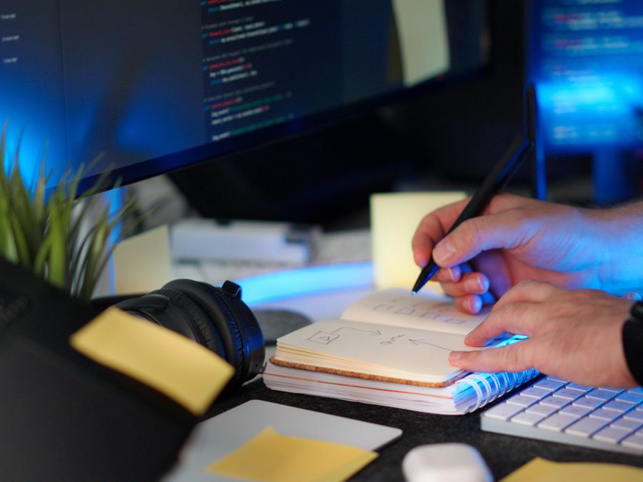 A person planning a software project with handwritten notes and code on a monitor in a modern workspace.