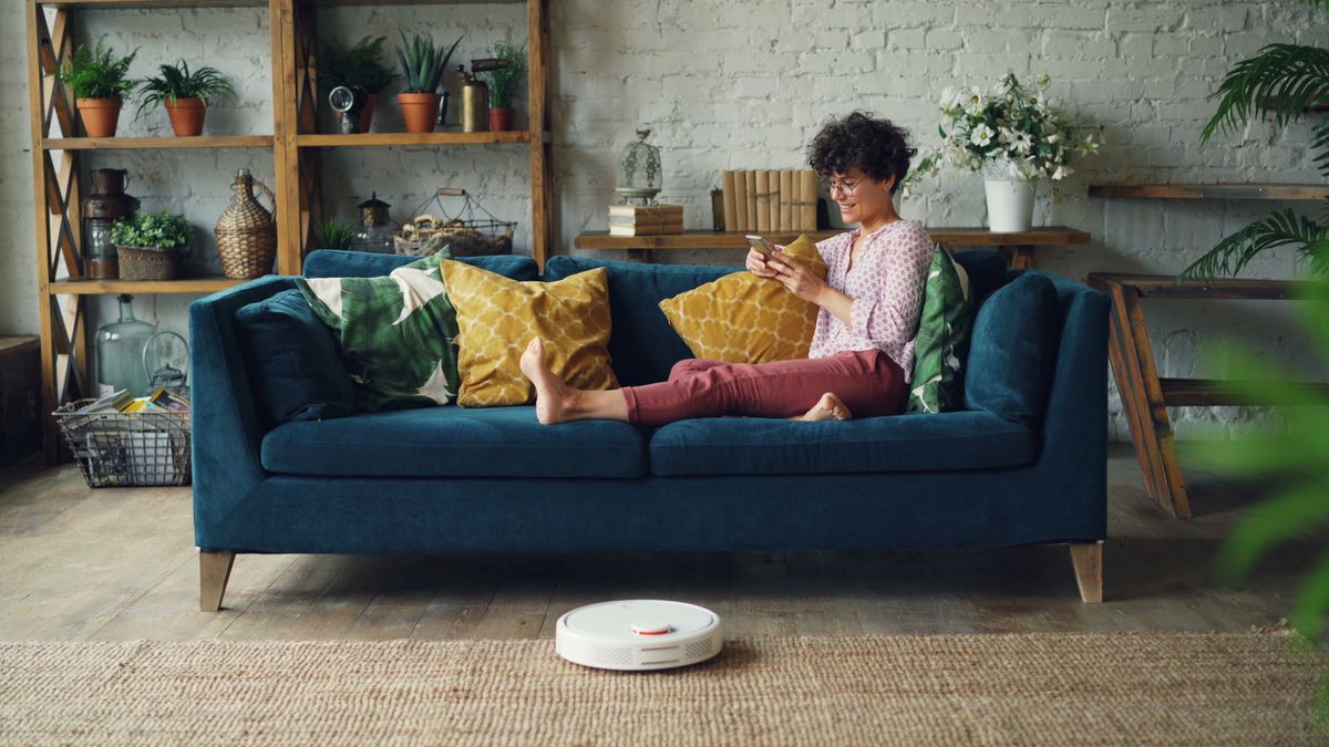 Woman relaxing on couch with phone as a robot vacuum cleans the floor.