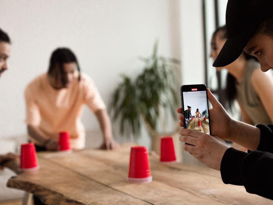 Group of young adults engaging in a creative game with red cups indoors, capturing moments on a smartphone.