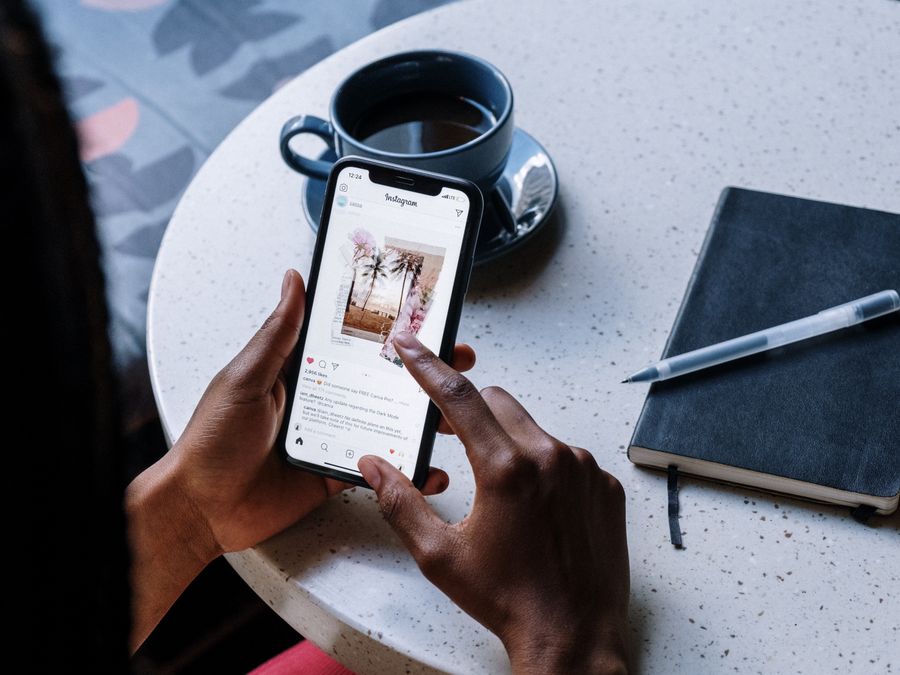 Hands holding smartphone at a cafe table with coffee and a notebook, focusing on social media browsing.