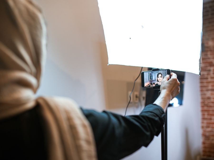A woman wearing a headscarf is recording a vlog with a smartphone and bright studio light.