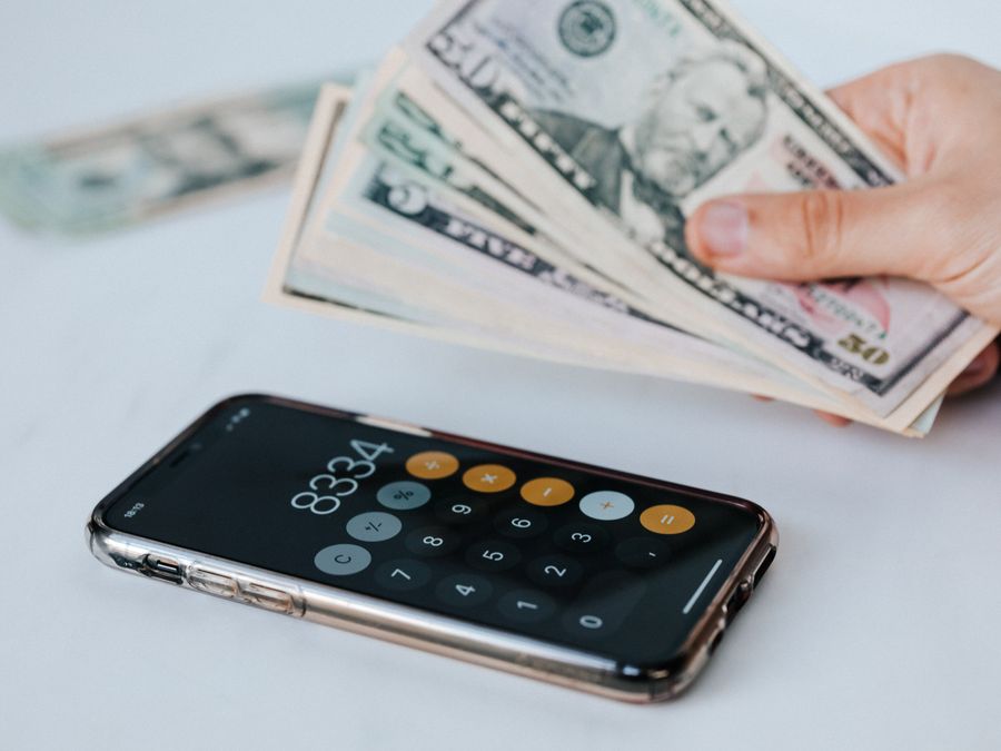 Calculator application on mobile phone screen on white surface near crop anonymous person holding pile of dollars