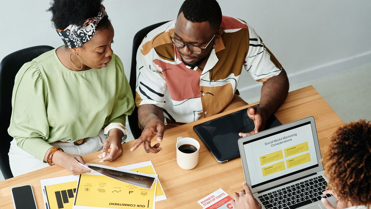 Three colleagues collaborate over marketing documents and a laptop in a modern office setting.