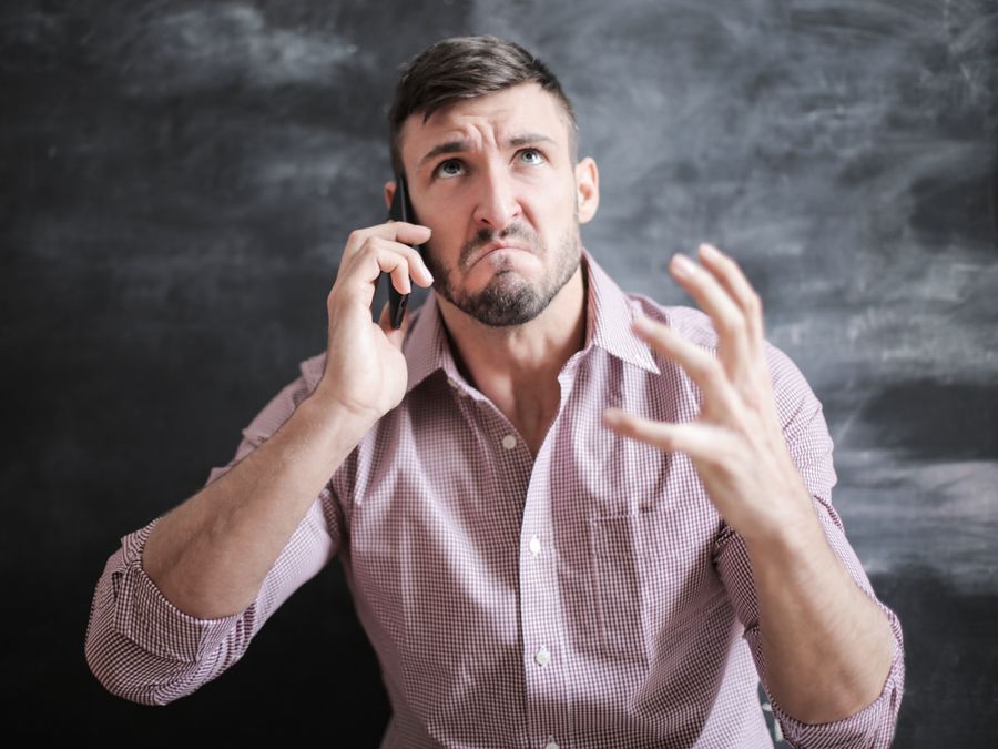 Adult man in pink dress shirt looking stressed while talking on a smartphone indoors.