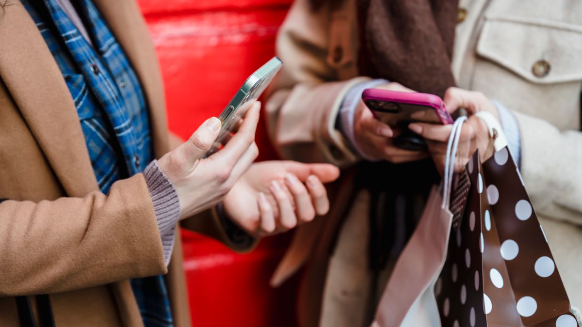 Crop faceless female friends in stylish outfits using smartphones while standing on street near red wall with shopping bags