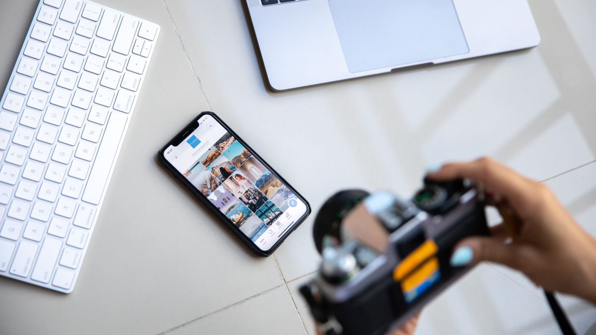 Person using vintage camera with smartphone and laptop in creative setup.