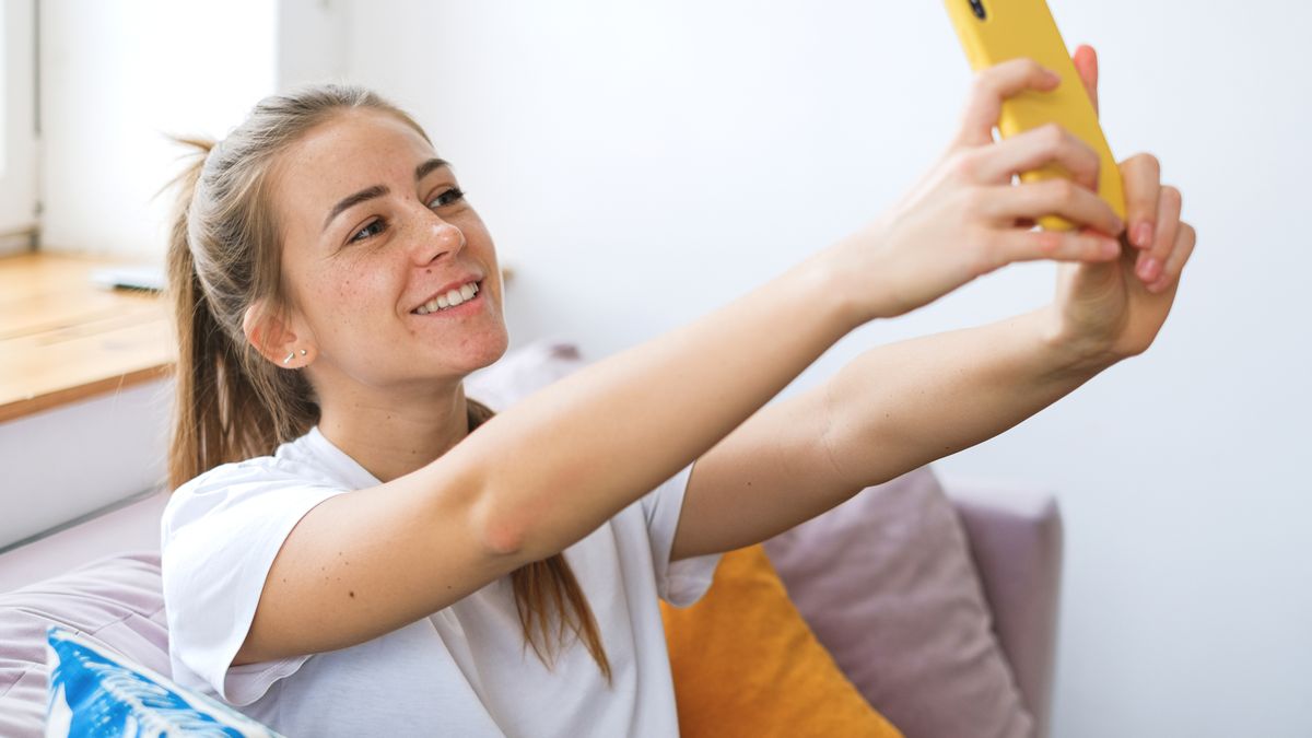 Smiling woman taking a selfie indoors with a yellow phone, relaxed at home.