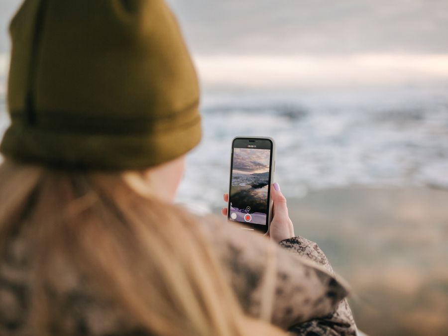 A person with a beanie using a smartphone to film the ocean view.